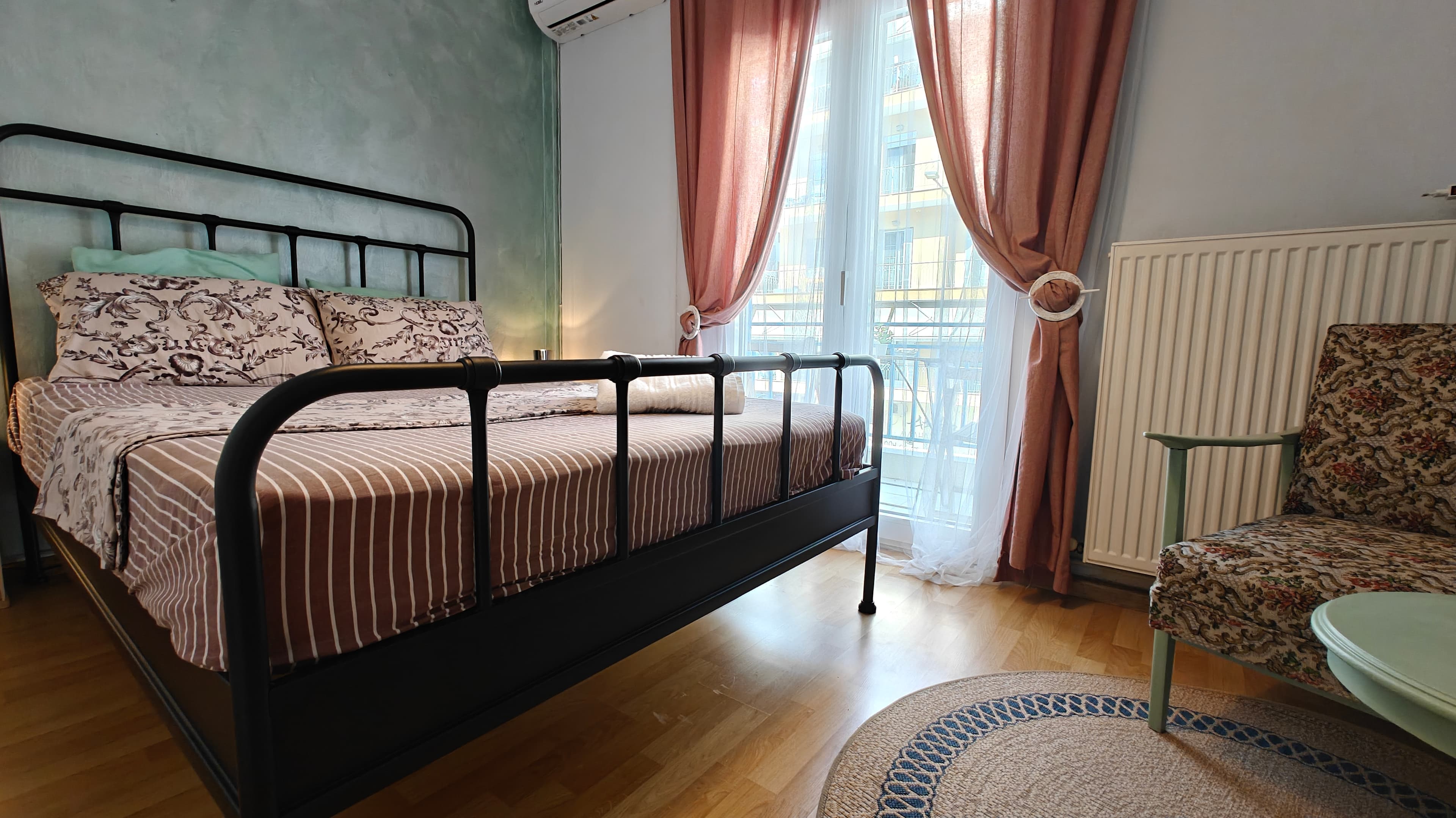 Black metal bed with striped bedding next to pink curtains and a floral armchair.