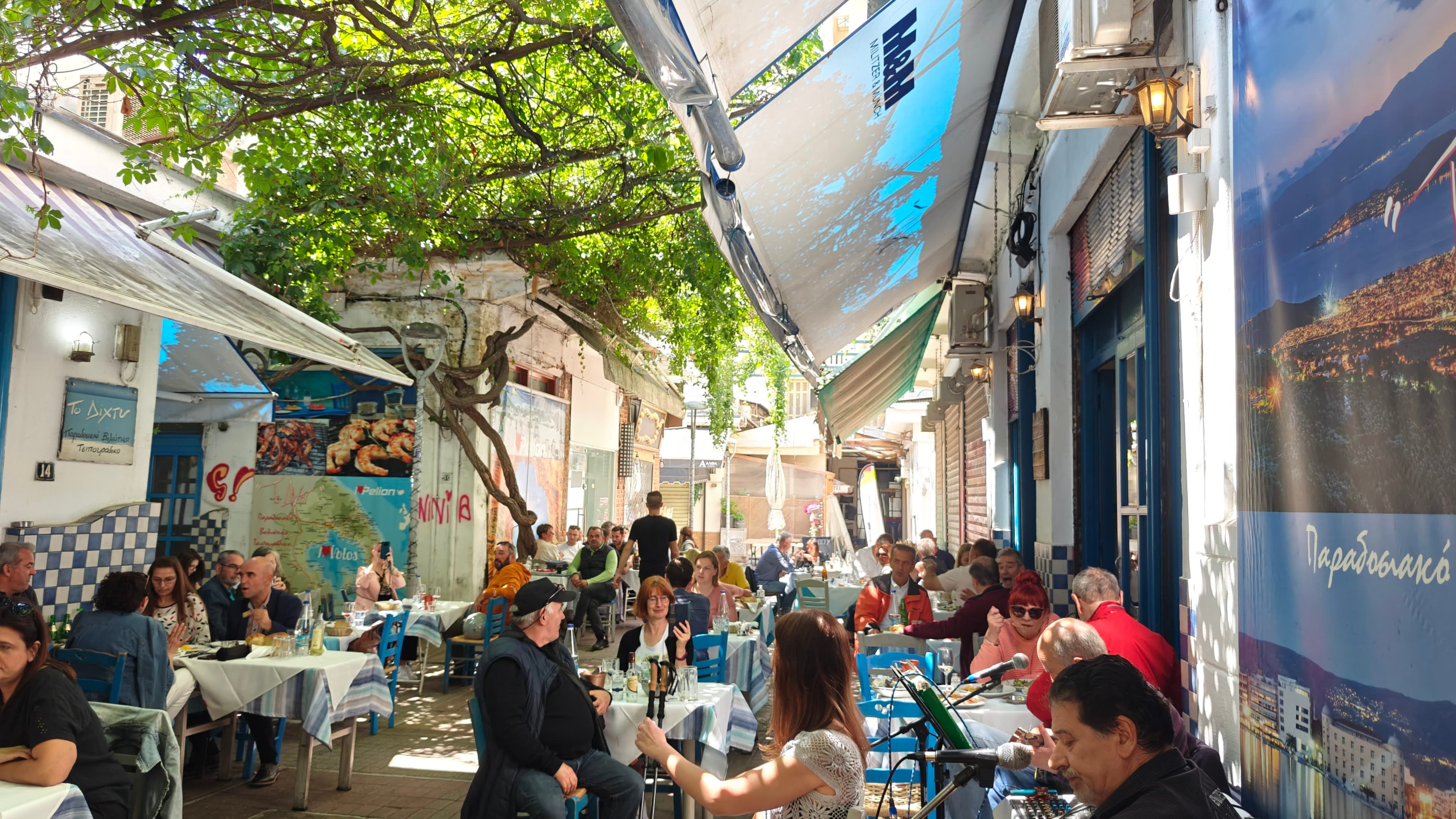 Crowded outdoor cafe in a narrow Greek alleyway shaded by a lush tree canopy.