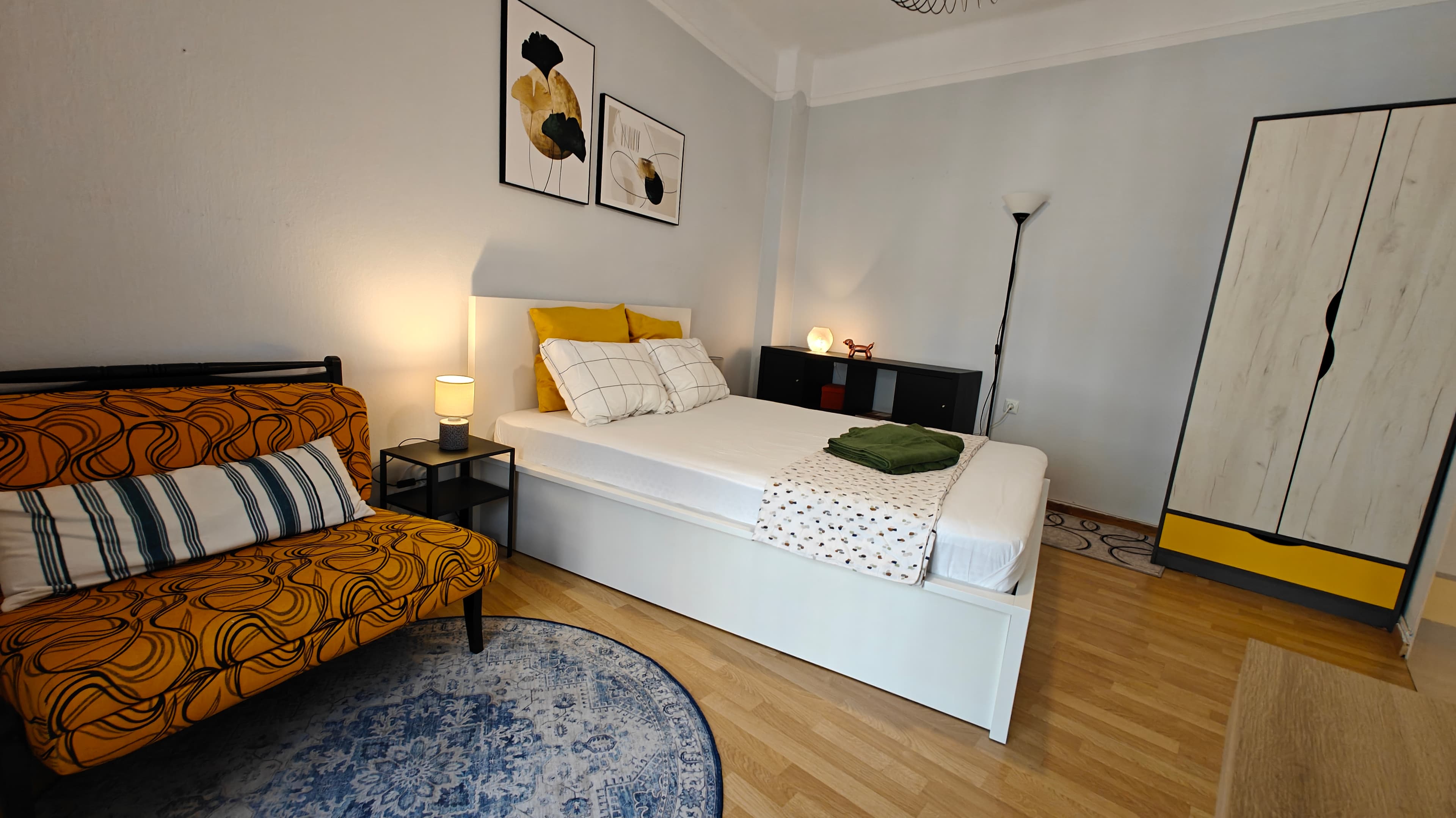 Modern bedroom with a white bed, yellow patterned sofa, blue rug, and framed wall art.